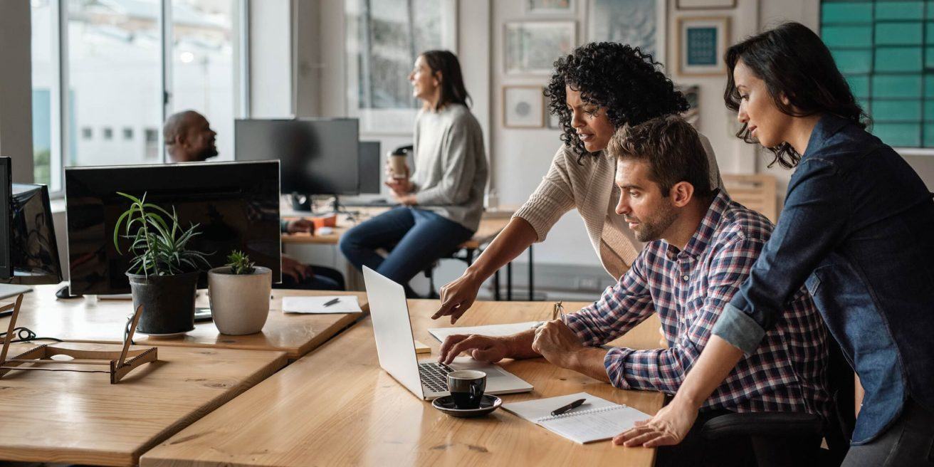 two people looking at a laptop screen and one person working in the background